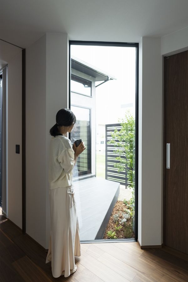マグカップを両手で持って窓辺に立ち、庭を眺める女性